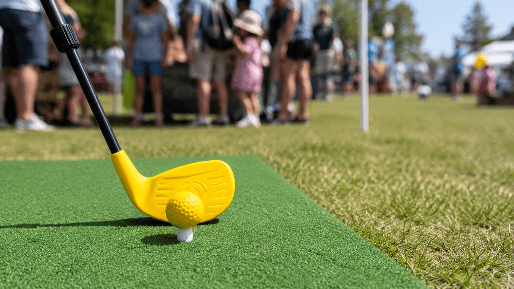 Yellow golf club and ball on a green putting surface with spectators in the background. Bananas Golf - Raleigh, NC - Triangle Area - Nerf Golf Hole In One Nerf Golf Challenge 